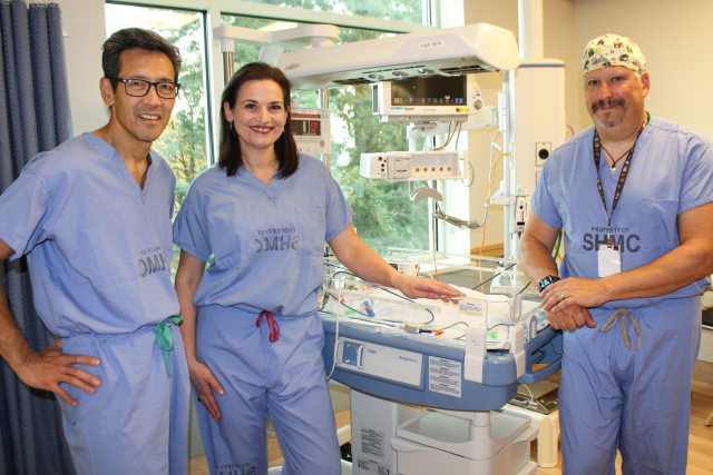 Three smiling PeaceHealth physicians stand next to a bed in the NICU