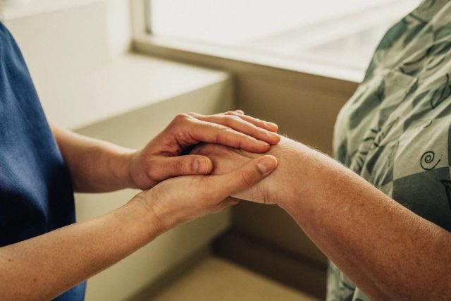A caregiver holds the hand of a patient in a hospital gown.