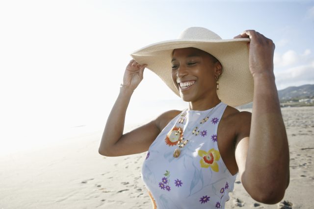 Black woman adjusts sun hat as she walks on sandy beach