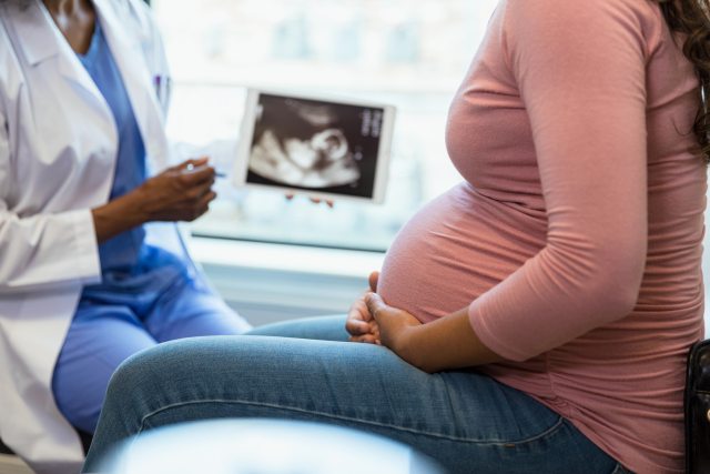 Photo of a pregnant woman with her hands on her belly in the foreground and her doctor with an ultrasound on a digital tablet in the background.