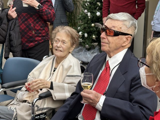 An elderly couple sits side by side at a reception. They wear formal clothing and hold glasses of cider in their hands.
