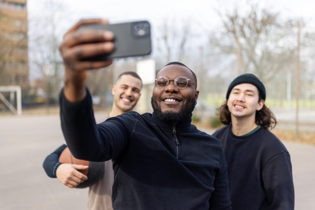 Black man wearing glasses uses cell phone to take selfie with 2 other men on outdoor basketball court