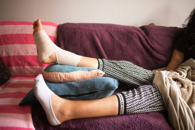 A young person on a couch puts wrapped ankle and foot on a pile of pillows
