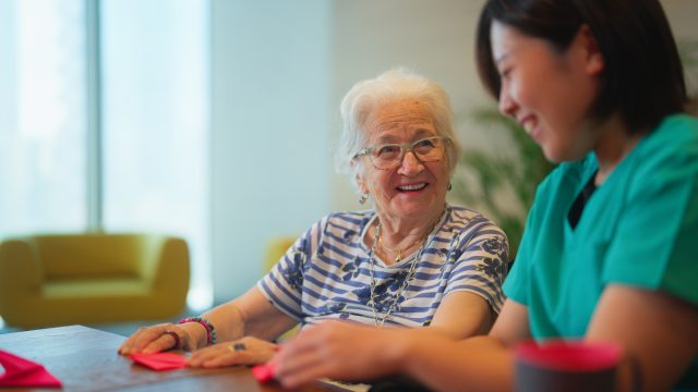 Older woman smiles at a young health caregiver as they fold paper at a table