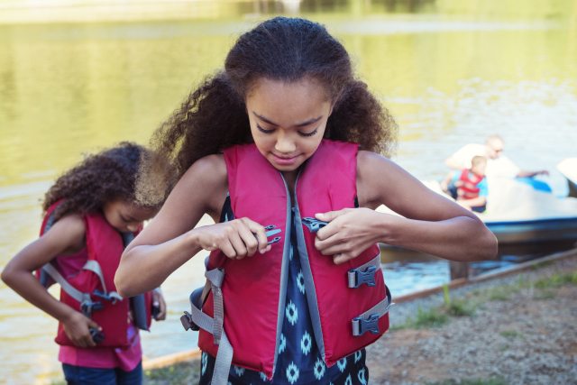 Young Black girls putting on bright pink life vests by the shore of a lake