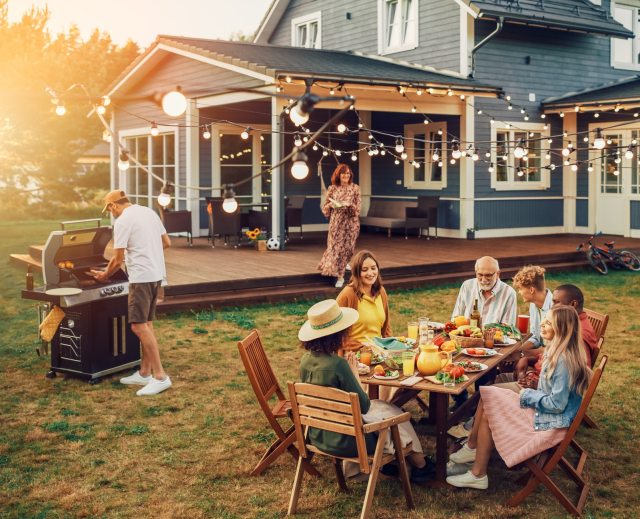 A summer barbecue in front of a blue hourse and deck with family & friends gathered around a table