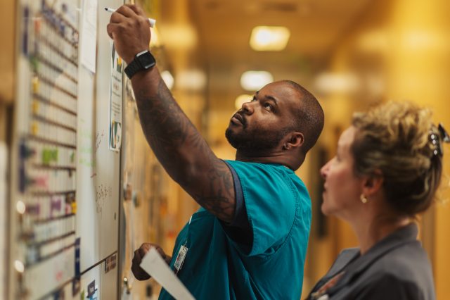 A nurse writes on a clinical whiteboard as another nurse watches and writes in a chart