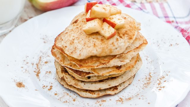 White plate with a stack of apple cinnamon pancakes. There is also a cup of milk and a fork on the table.