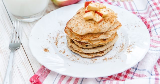 White plate with a stack of apple cinnamon pancakes. There is also a cup of milk and a fork on the table.