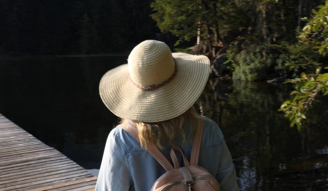 A blonde woman wearing a wide-brim sun hat and a backpack walks away from the camera along a dock at the lake 