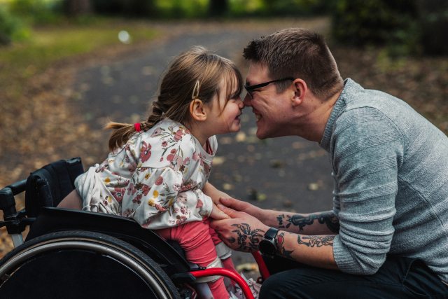 A man smiling and facing a young girl in a wheelchair, also smiling, on a nature path