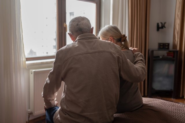 An older couple faces away from the camera to look out their bedroom window