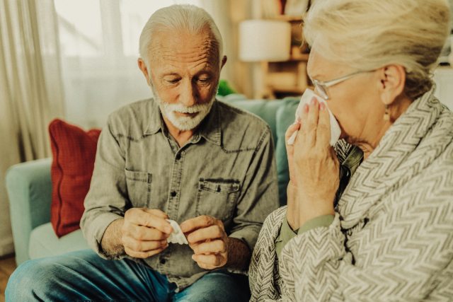 man opens medication packet near woman using tissue on nose