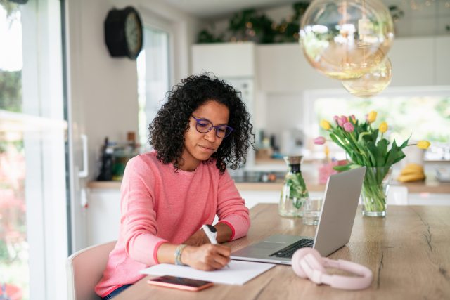 Woman wearing glasses sits at kitchen table, writing with a pen next to laptop