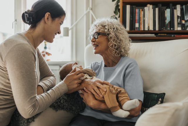 Grandmotherly woman holds baby while smiling at young mother