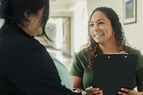 A caregiver holding a clipboard smiles while speaking with a person in the foreground