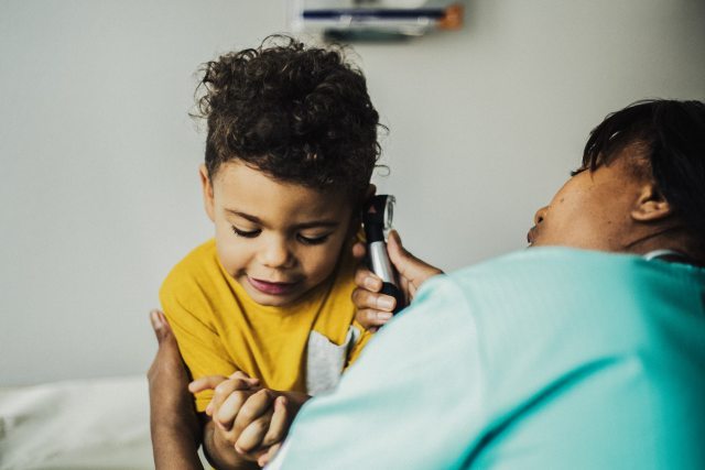 A young patient sits in a clinical setting while a pediatrician examines their ear with an otoscope
