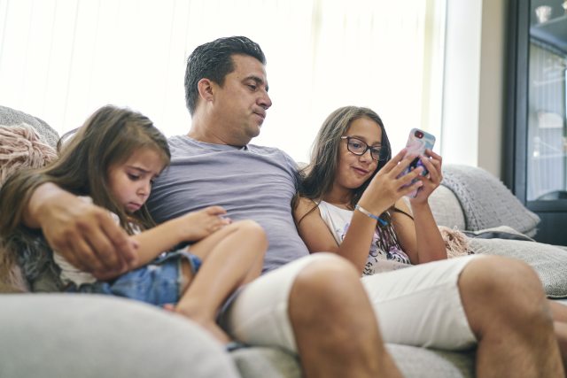 Father sits on couch looking at phone screen with young daughters