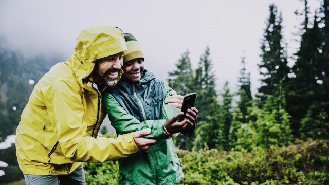 A man and boy in rain jackets take a selfie while on a hike in the rainy forest.