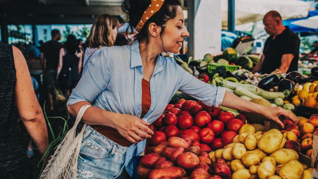 A woman at a farmer's market reaches over a vegetable stand to grab a potato