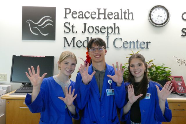 Three PeaceHealth Volunteers standing in front of the St. Joseph Medical Center sign, posing, with their hands up
