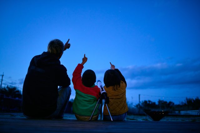 adult and two children sit outside and point at the darkening sky
