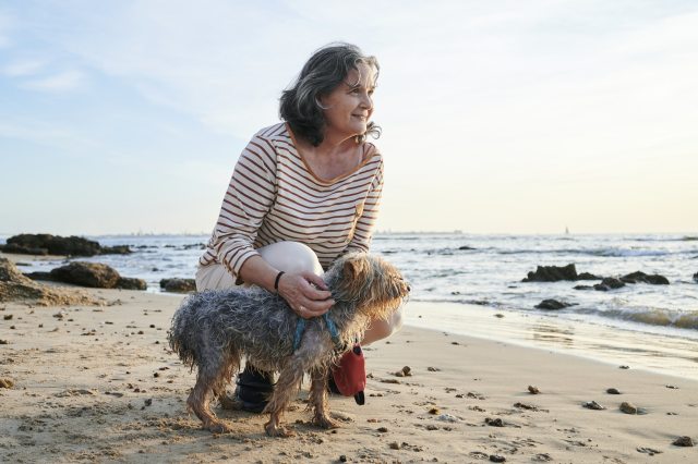 Woman crouches by her small dog on the beach