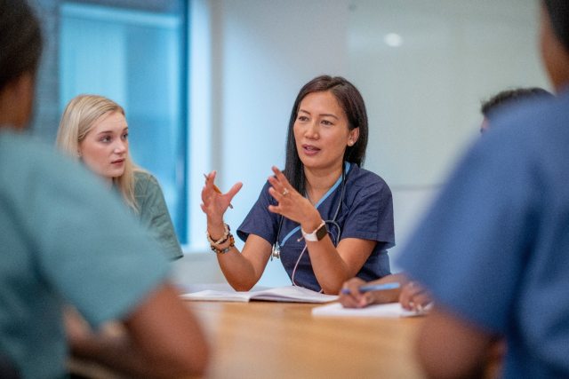 A woman caregiver sitting at a table and speaking at a meeting. Other caregivers are partially in frame but out of focus