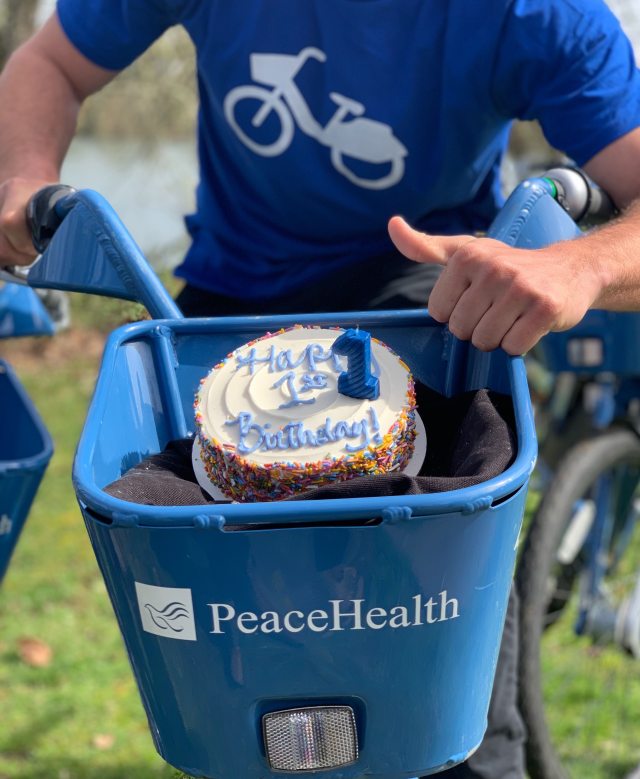 A PeaceHealth employee riding a bicycle with a cake in the basket, spelling out "Happy 1st Birthday"