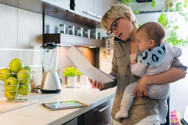A busy mother on her phone, holds her baby in the kitchen while looking at a piece of paper