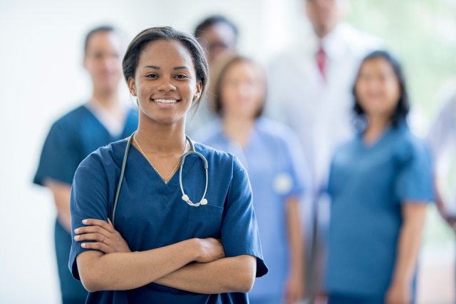 A smiling nurse stands in front of a group of other nurses, blurred in the background