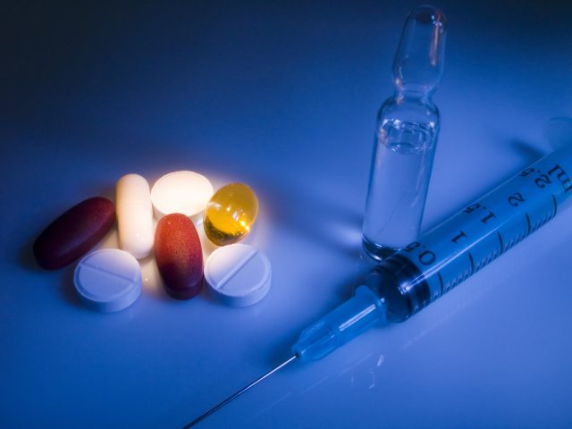 Dark image of a syringe and glass tube on the right with various colored pills highlighted to the left