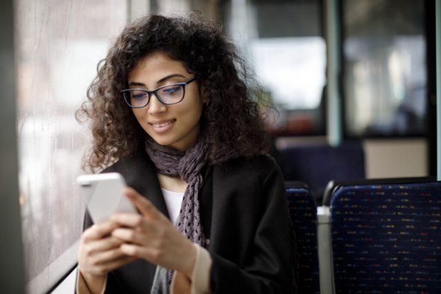 A young woman sitting on the bus or train, checking her mobile phone and smiling