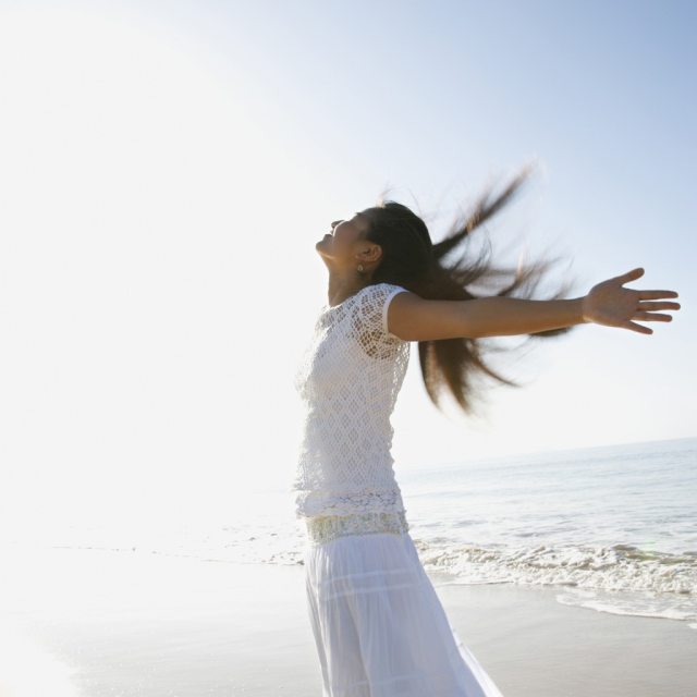 A woman in white stands at the beach with her arms outstretched and face towards the sun