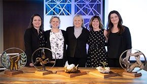 A group of five smiling women standing at a table with awards