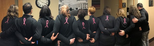 A group of people in black shirts with a pink ribbon on the back embrace in a large circle with their backs to the camera