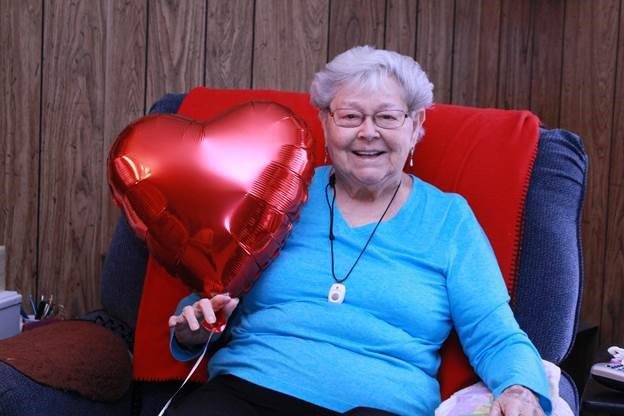 An elderly woman smiles while sitting and holding a heart shaped balloon