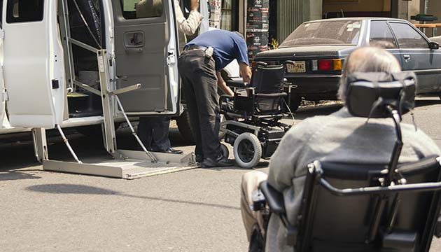 A man in a motorized wheelchair rolls up to another person adjusting a power wheelchair next to van