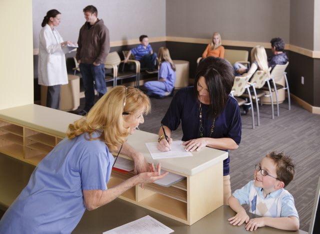 A nurse speaks with young patient while the patient's mother fills in patient forms in an office setting
