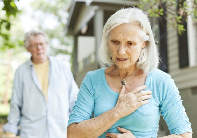 Older woman facing the camera puts her hand to her chest in sudden pain with older man in background looking concerned