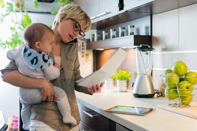 A woman holding a baby while in the kitchen looks at a piece of paper and talks on her mobile phone