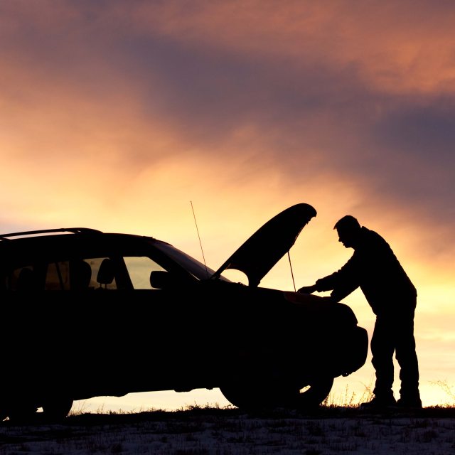 Silhouette of a man and car with the hood up, against a sunset of purple and pink hued sky