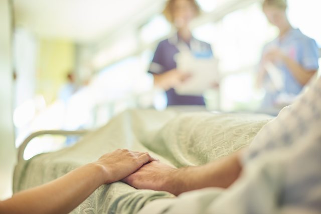 Close-up of a person holding a patient's hand with healthcare providers blurred in the background
