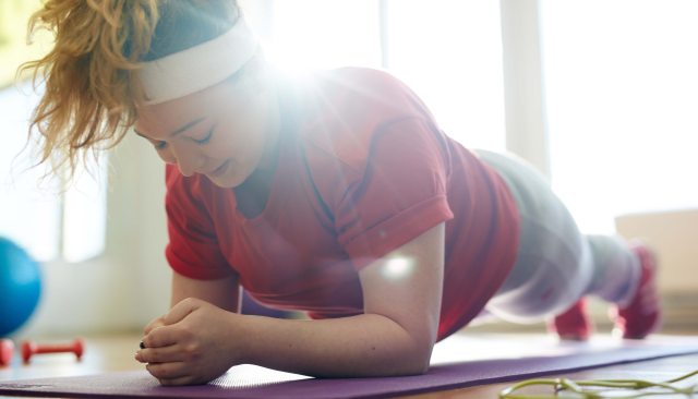 A woman performs a plank exercise on a yoga mat with sunlight beaming in from the background