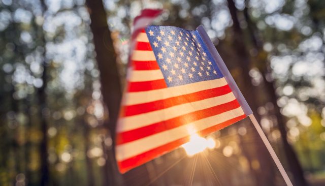 A United States flag waves in front of sunlight beaming through trees
